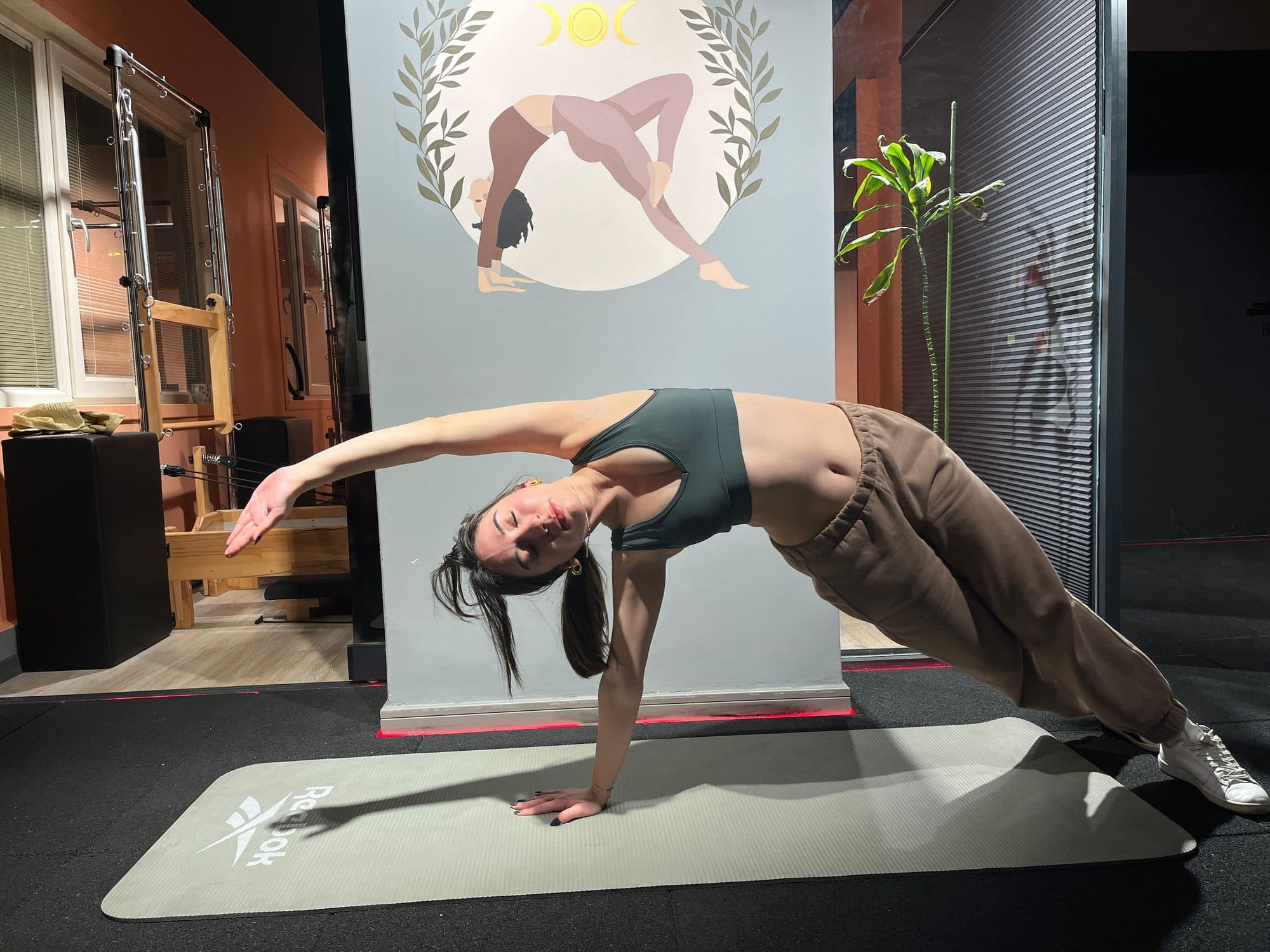 Gözleri kapalı halde tek kolla yan köprü pozisyonunda gövdesini esneten kadın, arkada pilates stüdyosu